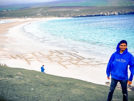 Man in blue hoodie standing on a grassy hill overlooking a beach with "SHETLAND FOSSIL FREE" written in the sand, with a child and cliffs in the background.