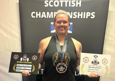 A person smiling, holding two world record certificates and wearing a medal at the scottish championships event.