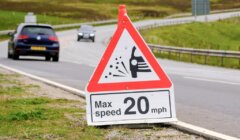 A triangular sign warning of loose gravel on the road with a maximum speed limit of 20 mph is positioned beside a rural road. Two cars are visible traveling on the road in the background.