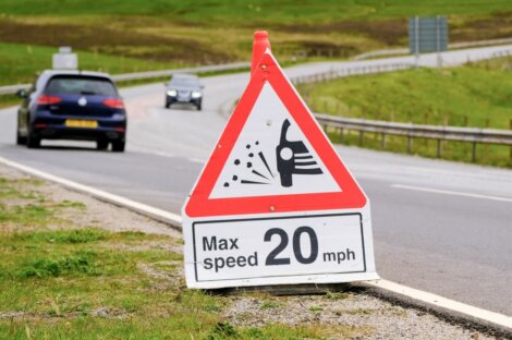 A triangular sign warning of loose gravel on the road with a maximum speed limit of 20 mph is positioned beside a rural road. Two cars are visible traveling on the road in the background.