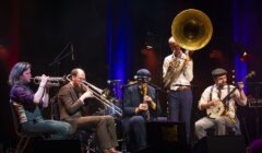 A jazz band performs live on stage, featuring musicians playing a trumpet, saxophone, trombone, and a sousaphone under colorful stage lighting.