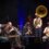 A jazz band performs live on stage, featuring musicians playing a trumpet, saxophone, trombone, and a sousaphone under colorful stage lighting.