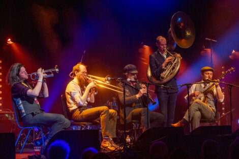 Four musicians perform on stage, playing trumpet, trombone, sousaphone, and saxophone under vibrant stage lighting.