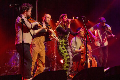 A folk band with five members playing violin, flute, and guitar on a dimly lit stage during a live concert.