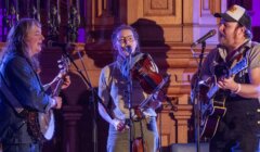 Three musicians perform in an ornate hall, playing a guitar, a violin, and singing into microphones.