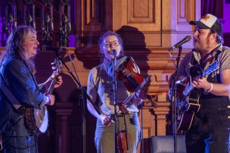 Three musicians perform in an ornate hall, playing a guitar, a violin, and singing into microphones.