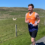 A man in an orange and blue sports vest is jogging on a rural road with green fields, sheep, and a house in the background under a clear blue sky.