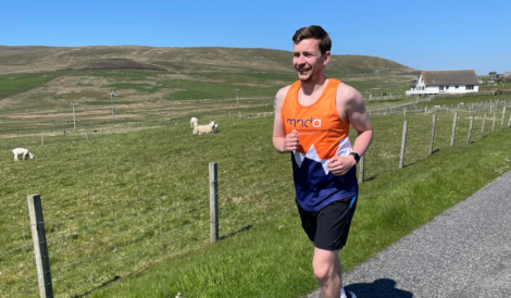 A man in an orange and blue sports vest is jogging on a rural road with green fields, sheep, and a house in the background under a clear blue sky.