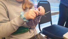 A person wearing a light grey sweater is knitting a purple and white patterned item while sitting next to a window, with empty chairs visible beside them.