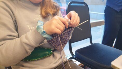 A person wearing a light grey sweater is knitting a purple and white patterned item while sitting next to a window, with empty chairs visible beside them.