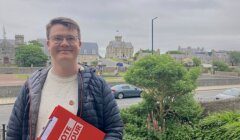 A person stands outdoors holding a clipboard with "VOTE LABOUR" written on it. Buildings and greenery are visible in the background.