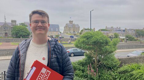 A person stands outdoors holding a clipboard with "VOTE LABOUR" written on it. Buildings and greenery are visible in the background.