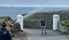 A firefighter directs a hose to spray water on a smiling man standing on a path near stone walls and overlooking the ocean. Others observe in the background.