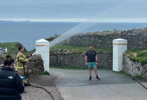 A firefighter directs a hose to spray water on a smiling man standing on a path near stone walls and overlooking the ocean. Others observe in the background.