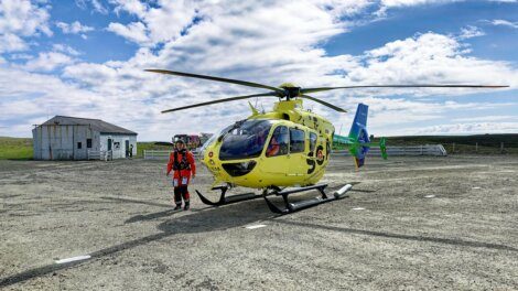 A yellow helicopter is parked on a gravel surface, with a person in red gear standing next to it. An old building and a cloudy sky are in the background.