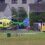 A yellow and green SGAA helicopter lands near an ambulance outside residential buildings.