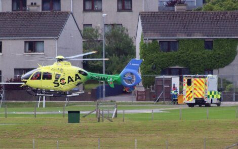 A yellow and green SGAA helicopter lands near an ambulance outside residential buildings.