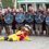 A group of people dressed as Vikings with shields and axes pose for a photo behind a person in a yellow and red mascot costume lying on the ground.