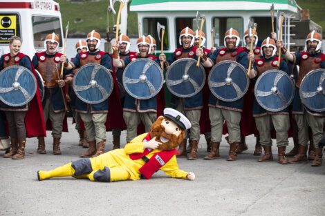 A group of people dressed as Vikings with shields and axes pose for a photo behind a person in a yellow and red mascot costume lying on the ground.