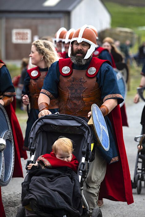 A group of people dressed as Viking warriors, including a man pushing a stroller with a toddler. They are walking outdoors with buildings and green lawns in the background.