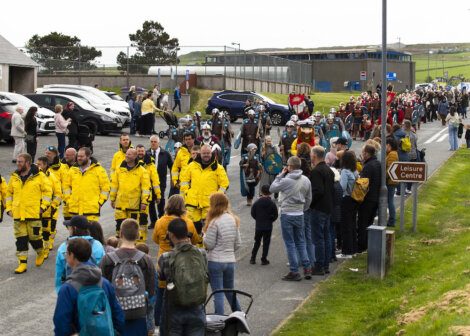 A crowd watches as a procession, with people in yellow jackets and some in medieval costumes, goes past a parking lot and towards a leisure center.