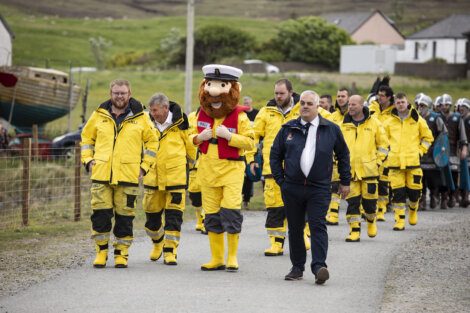 A group of people in yellow raincoats and boots, with a mascot in a captain's hat, walk behind a man in a dark jacket along a rural path by boats and houses.