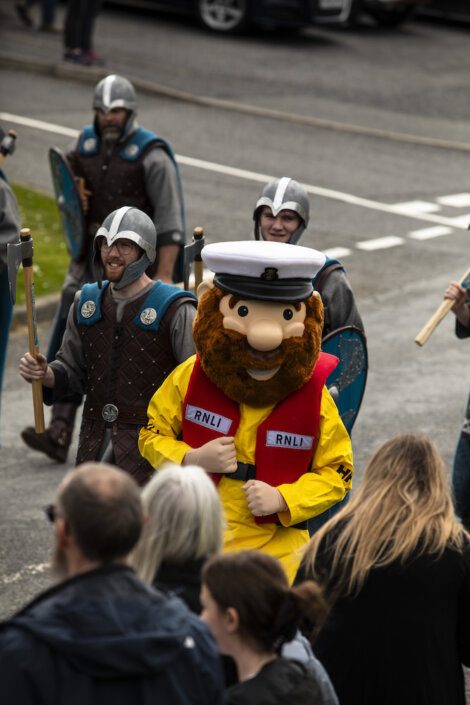 A person in a yellow RNLI uniform and large mascot head stands among people dressed in medieval costumes holding shields and spears, with an audience watching in the foreground.