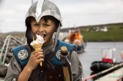 A boy in medieval armor eats a soft-serve ice cream cone topped with a chocolate flake, standing outdoors near a waterfront with boats and grassy land in the background.