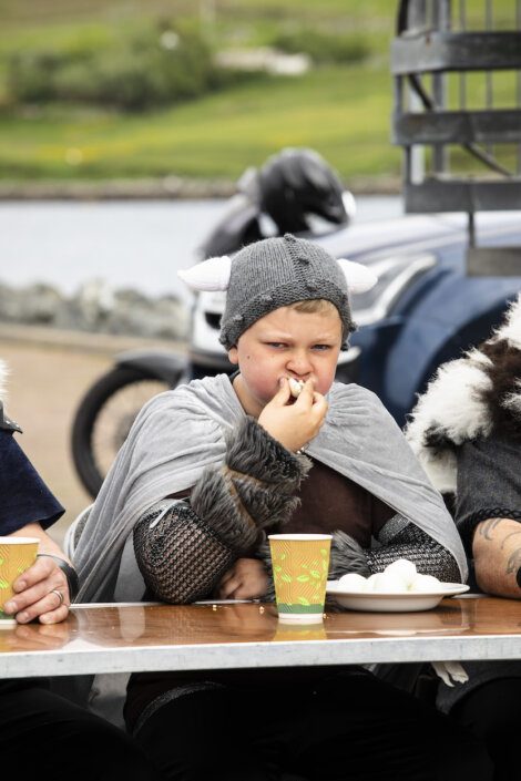 A child in a gray costume with a horned cap eats food at an outdoor table, with a cup and plate in front of them. A blurred background shows greenery and a parked motorcycle.