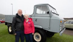 Two elderly individuals stand together outdoors beside a vintage gray truck. The man is wearing a black jacket, and the woman is wearing a red jacket. Other cars are visible in the background.