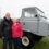 Two elderly individuals stand together outdoors beside a vintage gray truck. The man is wearing a black jacket, and the woman is wearing a red jacket. Other cars are visible in the background.