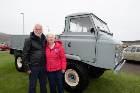 Two elderly individuals stand together outdoors beside a vintage gray truck. The man is wearing a black jacket, and the woman is wearing a red jacket. Other cars are visible in the background.