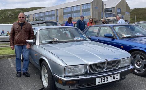 A man in a brown leather jacket stands next to a silver Jaguar car with the license plate J199 TAC at a car show. Other people and cars are visible in the background.