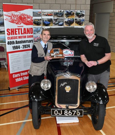 Two men pose with a trophy in front of a vintage black Austin car at the Shetland Classic Motor Show. A banner behind them mentions the show's 40th anniversary from 1984 to 2024.