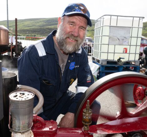 A man wearing a blue cap and overalls smiles while standing next to a spinning engine outdoors, with green hills in the background.