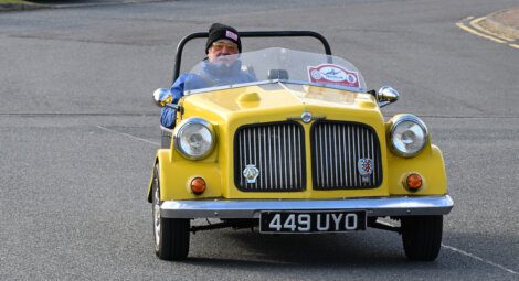 A person drives a small, yellow vintage car with a black license plate that reads "449 UYO" on a road. The driver is wearing a hat and sunglasses.