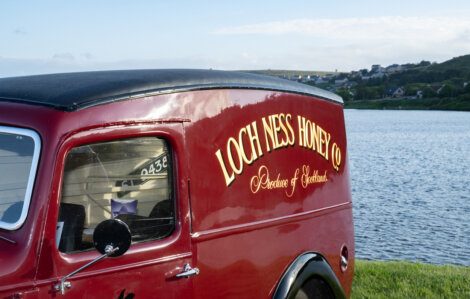 A red van with "Loch Ness Honey Co. Produce of Scotland" painted on the side, parked near a body of water with a village in the background.