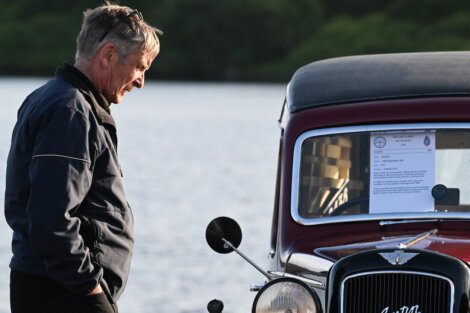 Man in a dark jacket stands next to a vintage car by the water, examining a document displayed on the windshield of the vehicle.