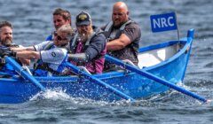 Four men are rowing a blue boat labeled "NRC" in the water. They are wearing life jackets and are focused on rowing.