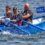 Four men are rowing a blue boat labeled "NRC" in the water. They are wearing life jackets and are focused on rowing.