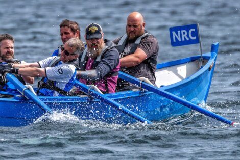 Four men are rowing a blue boat labeled "NRC" in the water. They are wearing life jackets and are focused on rowing.