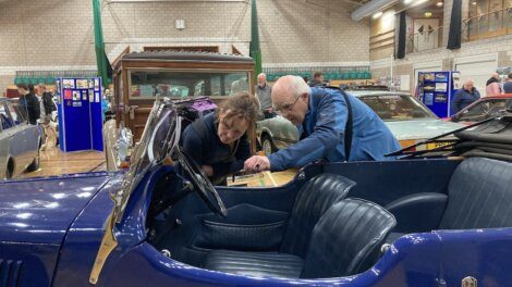 Two people examine the interior of a vintage blue convertible car at an indoor car exhibition. Other classic cars and attendees are visible in the background.