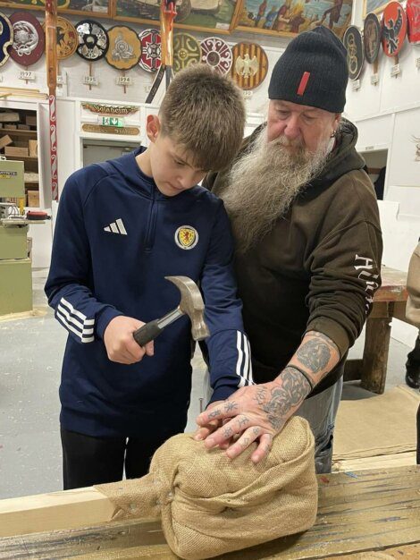 A young boy in a blue tracksuit uses a hammer on a wooden plank under the supervision of an older man with tattoos and a beanie in a workshop.