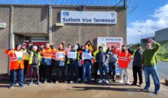 A group of workers wearing high-visibility jackets and holding Unite union posters stand outside a building labeled "Sullom Voe Terminal," with barbed wire visible above the entrance.