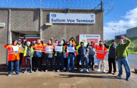 A group of workers wearing high-visibility jackets and holding Unite union posters stand outside a building labeled "Sullom Voe Terminal," with barbed wire visible above the entrance.