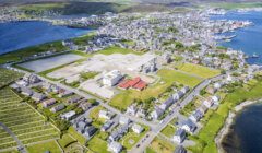 Aerial view of a coastal town with residential buildings, a cemetery, green spaces, and large construction areas near the shoreline and harbor.