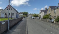 A small road on a sunny day with a 20 mph speed limit sign, a row of houses on both sides, and two cars parked along the curb.