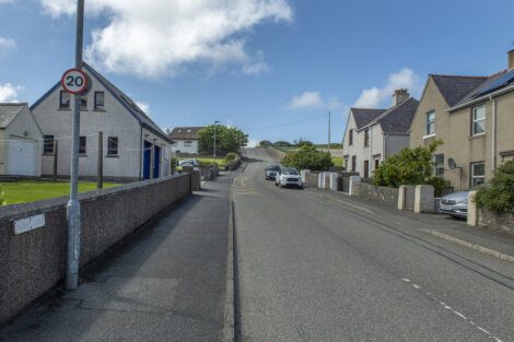 A small road on a sunny day with a 20 mph speed limit sign, a row of houses on both sides, and two cars parked along the curb.