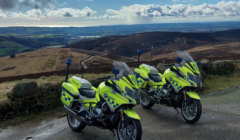 Two police motorcycles in high-visibility yellow parked on a scenic rural hillside road with a vast landscape and cloudy sky in the background.