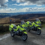 Two police motorcycles in high-visibility yellow parked on a scenic rural hillside road with a vast landscape and cloudy sky in the background.